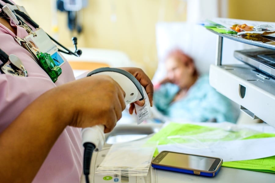 A nurse scans a barcode in a hospital room with a patient lying in a bed in the background